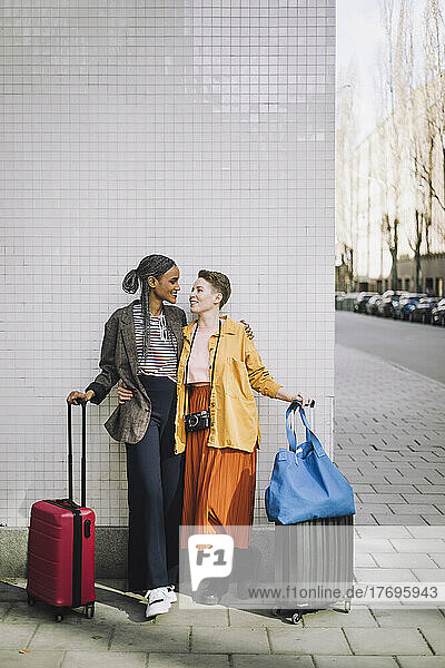 Lächelndes lesbisches Paar in voller Länge mit Gepäck auf Rädern an der Wand stehend