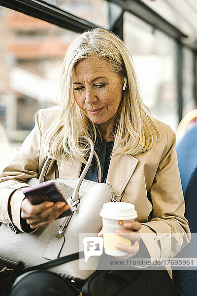 Weibliche Fachkraft  die ein Smartphone benutzt und einen Einwegbecher hält  während sie im Bus sitzt