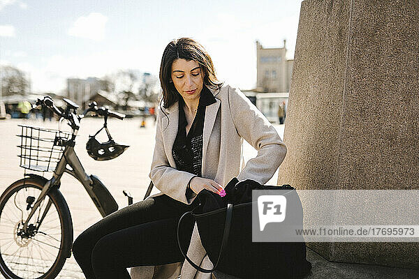 Businesswoman searching in purse while sitting by bicycle