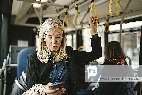 Businesswoman holding grab handle while using smart phone in bus