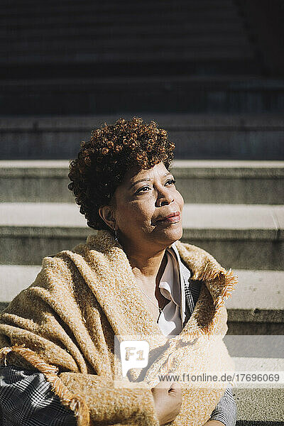 Nachdenkliche ältere Frau mit lockigem Haar  die an einem sonnigen Tag auf einer Treppe sitzt