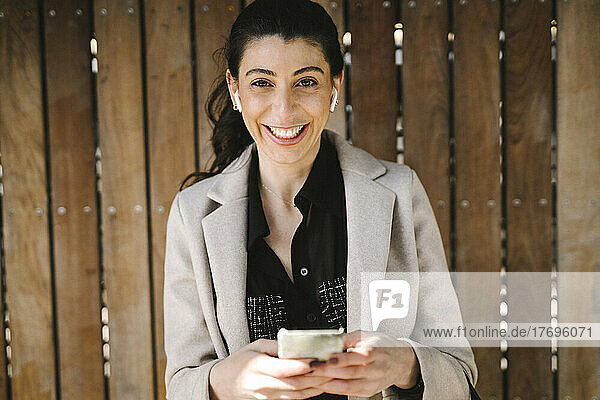 Porträt einer fröhlichen Geschäftsfrau  die ein Smartphone an eine Holzwand hält