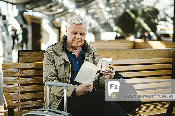 Senior businessman holding disposable cup reading book while sitting on bench at railroad station food court