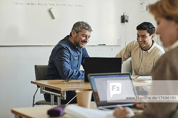 Smiling teacher and student looking at laptop while sitting in classroom