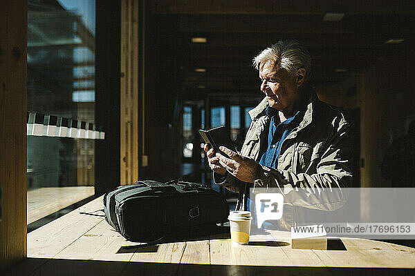 Geschäftsmann  der sein Smartphone benutzt  während er mit seinem Rucksack auf dem Tisch im Wartezimmer eines Bahnhofs steht