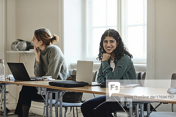 Portrait of smiling female student with laptop sitting by friend at desk in classroom