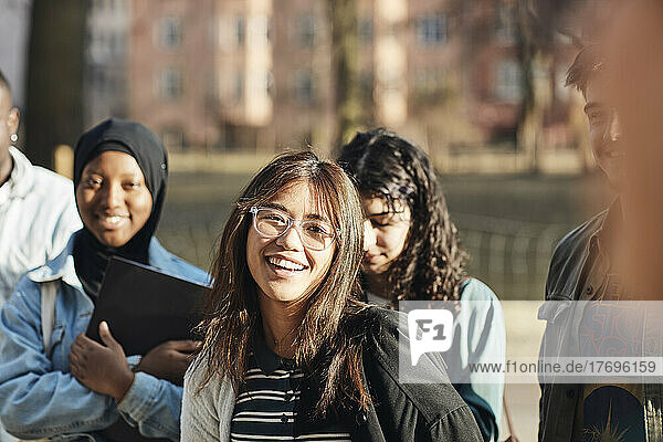 Portrait of smiling young female student with friends at university campus on sunny day