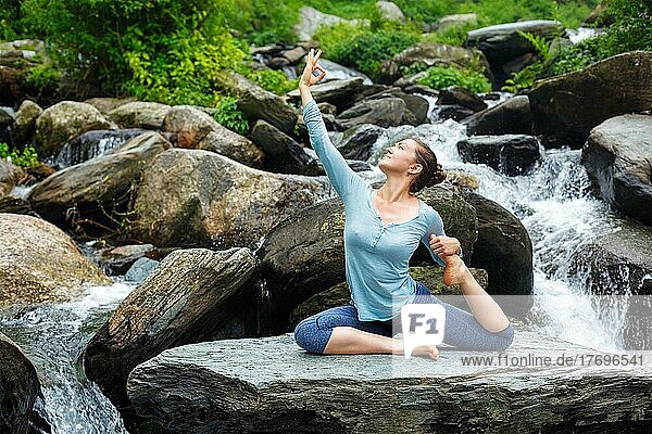 Young sporty fit woman doing yoga asana Eka pada rajakapotasana  one-legged king pigeon pose at tropical waterfall. Himachal Pradesh  India  Asia