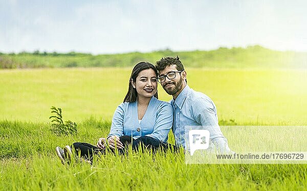 Junges verliebtes Paar  das im Gras sitzt und in die Kamera schaut  Zwei Liebende  die im Gras sitzen und in die Kamera schauen  Porträt eines verliebten Paares  das im Gras auf einem Feld sitzt