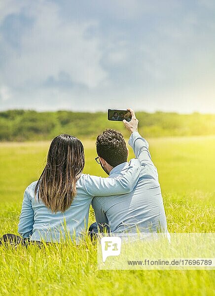 Verliebte  die mit ihrem Smartphone Selfies auf dem Feld machen  Lächelndes verliebtes Paar  das im Gras sitzt und Selfies macht  Junges verliebtes Paar  das ein Selfie auf dem Feld macht