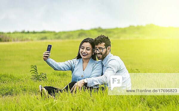 Lächelndes verliebtes Paar  das im Gras sitzt und Selfies macht  Junges verliebtes Paar  das ein Selfie auf dem Feld macht  Verliebte  die auf dem Feld Selfies mit ihrem Smartphone machen
