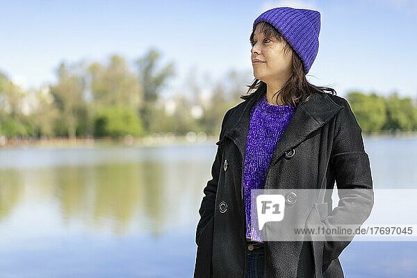Porträt einer jungen lateinischen Frau mit Pullover  Mantel und Wollmütze  die zur Seite auf einen See mit Kopierraum blickt