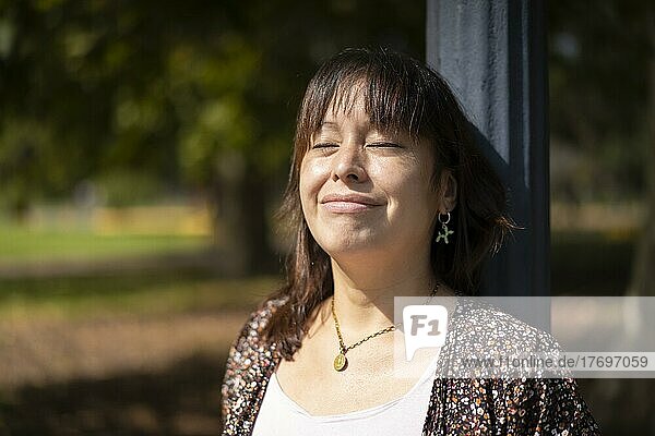 Porträt einer jungen lateinamerikanischen Frau in einem Park  die sich an einen Laternenpfahl lehnt und die Sonnenstrahlen auf ihrer Haut genießt  mit geschlossenen Augen