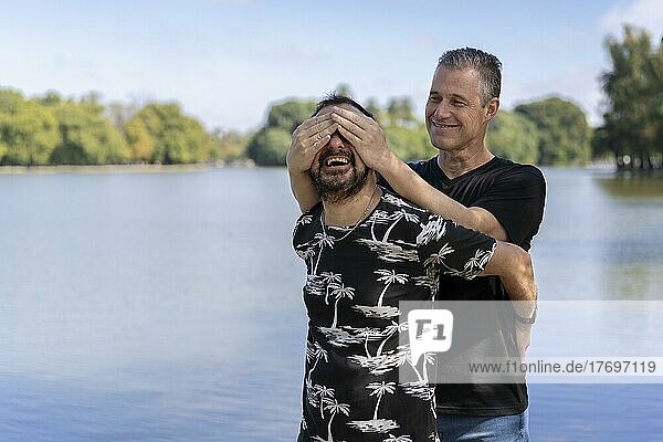 Couple of mature gay men  one surprising the other by covering his eyes in a lake. Concept of surprise  happiness  game