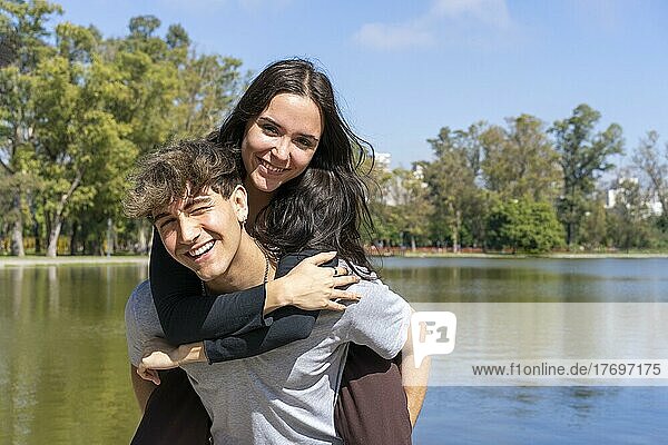 Junges Paar an einem See. Freund trägt seine Freundin auf dem Huckepack. Lustiger und glücklicher Ausdruck