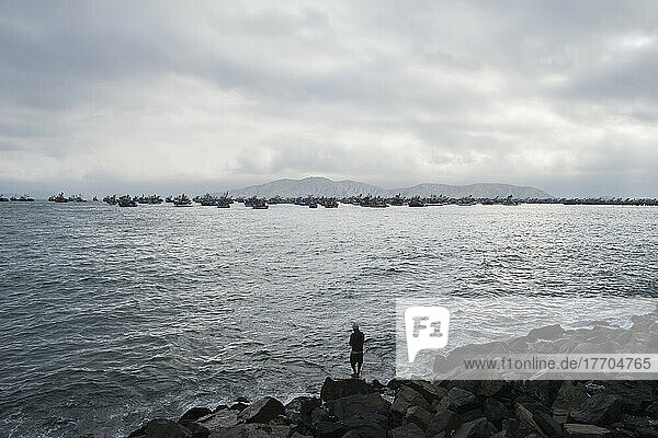 A Man Stands On A Rocky Shore Looking Out At Boats In A Busy Harbour; Chimbote  Peru