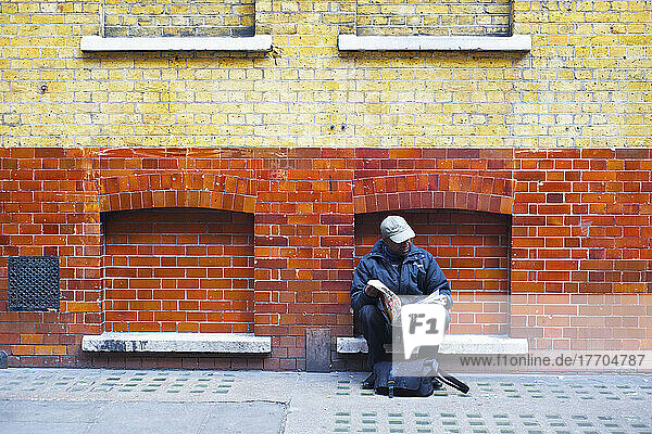 Ein Mann sitzt und liest eine Zeitung an einem Backsteingebäude; London  England