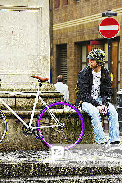Ein junger Mann sitzt auf einem Felsvorsprung neben einem Fahrrad; London  England