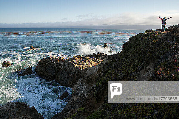 Eine Touristin auf einer Felsformation am Pazifischen Ozean. Als die Wellen an die Felsen schlagen  streckt sie begeistert ihre Arme aus; San Simeon Cove  San Simeon  Kalifornien