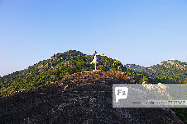 Eine junge Frau nimmt auf einem Hügel vor blauem Himmel eine Yogastellung ein; Ulpotha  Embogama  Sri Lanka