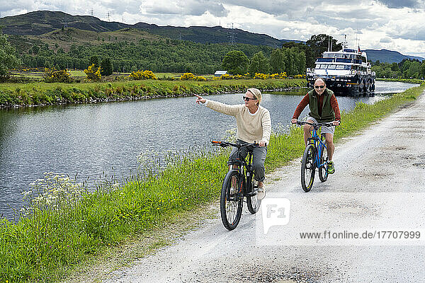 A man and woman ride bicycles along the Caledonian Canal tow path near Fort Augustus  Scotland. A Tour boat cruises along the canal in the background; Fort Augustus  Scotland