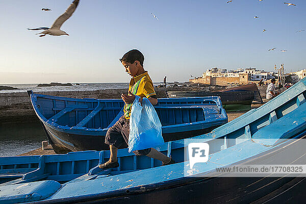 A young boy gingerly traverses a boat on the waterfront of Essaouira  Morocco; Essaouira  Morocco