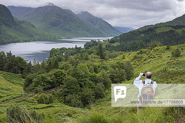 A hiker pauses along a trail to photograph Loch Shiel and the surrounding landscape at Glenfinnan  Scotland; Glenfinnan  Scotland
