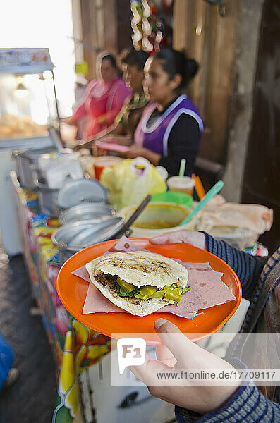 Frauenhände halten einen Teller mit einheimischem mexikanischem Essen  eine Gordita  neben dem Stand eines Lebensmittelverkäufers; San Luis De La Paz  Guanajuato  Mexiko