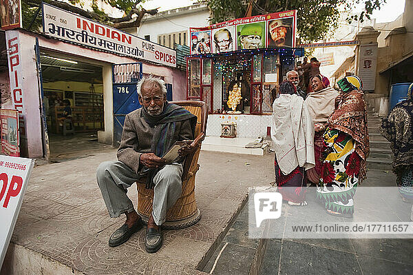Menschen stehen im Gespräch und ein Mann sitzt in einem Stuhl außerhalb von Einzelhandelsgeschäften; Udaipur  Indien