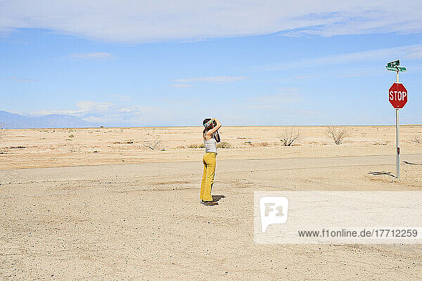 A young female photographer wearing colourful clothing stands photographing desert landscape in the town of Bombay Beach in the Sonoran Desert; Bombay Beach  California  United States of America