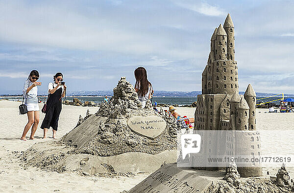 Two women taking a photograph of another woman standing next to a sandcastle creation on Coronado Beach; Coronado  California  United States of America