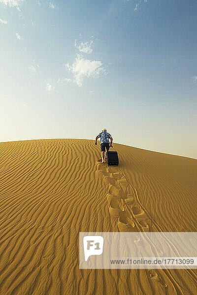 Barefoot Man With Suitcase Walking Up Sand Dune; Dubai  United Arab Emirates