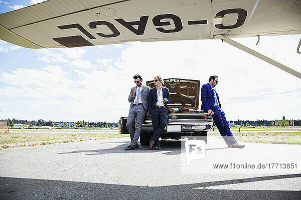 Three Businessmen Leaning Against A Car With An Open Trunk And An Airplane; Langley  British Columbia  Canada