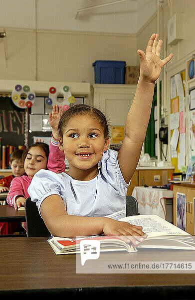 Six Year Old Girl Raising Her Hand At Her Desk In A Classroom