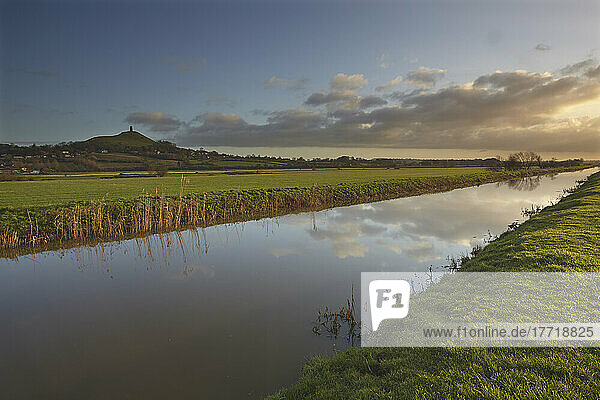 Das ruhige Wasser des Flusses Brue  in dem sich der Himmel spiegelt und das durch die Landschaft in der Nähe von Glastonbury fließt  mit Glastonbury Tor im Hintergrund  Somerset  Großbritannien; Somerset  England