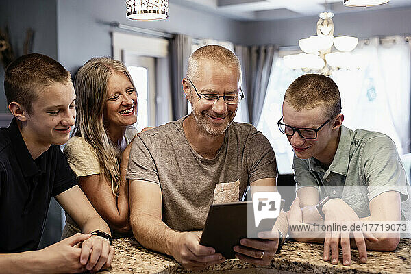 Parents and two sons sit at the kitchen island at home using a tablet and viewing content together; Edmonton  Alberta  Canada