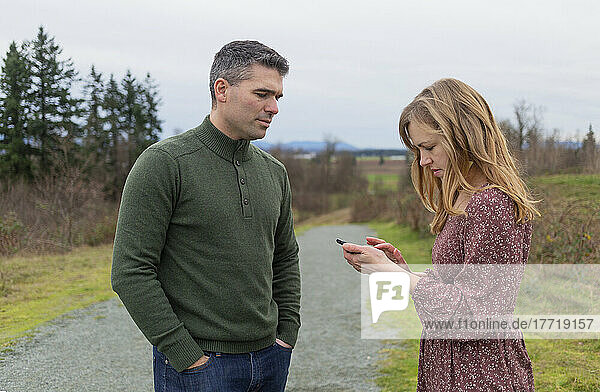 Paar im mittleren Erwachsenenalter steht in einem Park  die Frau benutzt ein Smartphone  während ihr Mann sie beobachtet; Aldergrove  British Columbia  Kanada