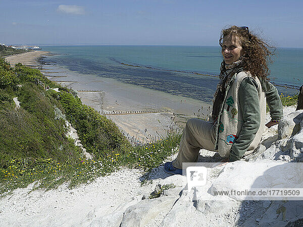 Woman sits on white rocks on a ridge above the beach and coastline looking back at the camera; Eastbourne  East Sussex  England