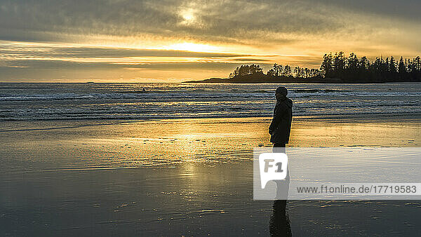 A woman stands on Long Beach at sunset looking out to the ocean  Pacific Rim National Park Reserve  Vancouver Island; British Columbia  Canada