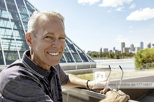 Outdoor portrait of senior man holding his eyeglasses  city skyline and modern architecture in the background; Edmonton  Alberta  Canada