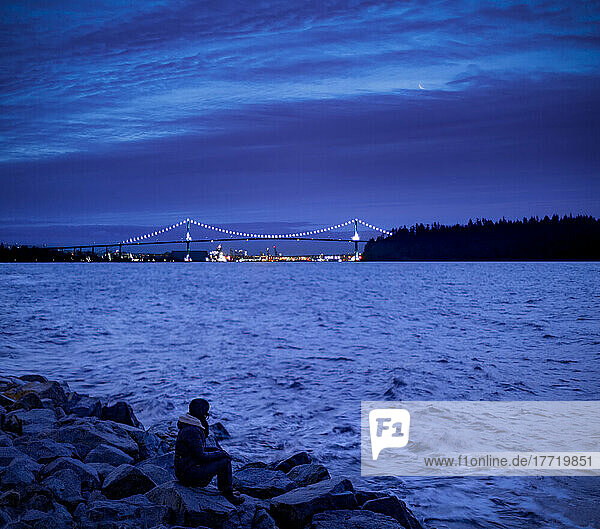 A woman sits on the rocks at the water's edge with views from the West Vancouver Seawall at twilight to the illuminated Lions Gate Bridge over Burrard Inlet in the distance; Vancouver  British Columbia  Canada