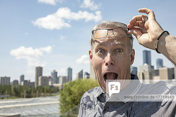 Ein älterer Mann steht im Freien und hebt seine Brille hoch  um mit einem überraschten Gesichtsausdruck in die Kamera zu schauen; Edmonton  Alberta  Kanada
