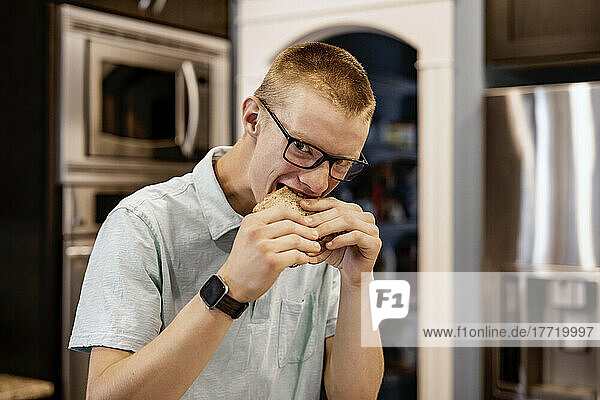 Junger Mann steht zu Hause in der Küche  isst ein Sandwich und schaut in die Kamera; Edmonton  Alberta  Kanada