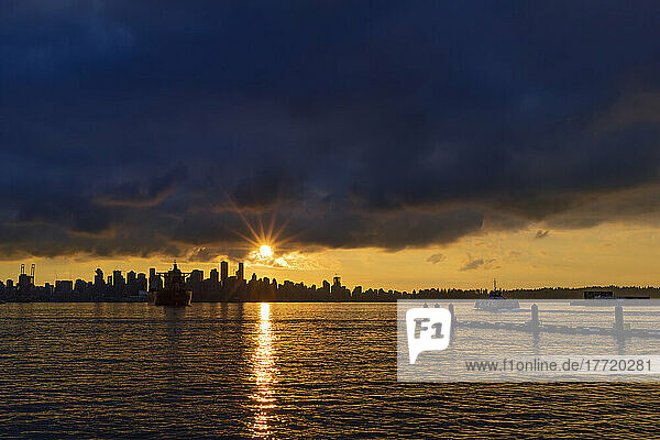 Goldener Sonnenuntergang hinter der Skyline der Innenstadt von Vancouver und dem Horizont mit Sonnenstrahlen  die sich auf dem Wasser des Burrard Inlet spiegeln; North Vancouver  British Columbia  Kanada