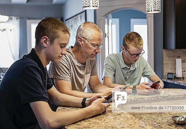 Father and two sons sit at the kitchen island at home using their smart phones; Edmonton  Alberta  Canada