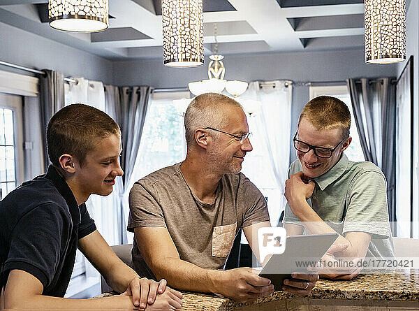 Vater und zwei Söhne sitzen zu Hause an der Kücheninsel  benutzen ein Tablet und reden miteinander; Edmonton  Alberta  Kanada