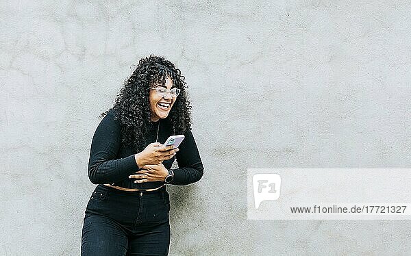Pretty girl happy with her cell phone  young latin woman happy with her cell phone leaning against a wall. A young latin woman laughing with her cell phone leaning against a wall