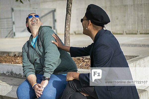 Ein schwules lateinamerikanisches Paar sitzt auf einer Bank in einem Park  trägt modische Hüte und Sonnenbrillen und sieht sich lachend an