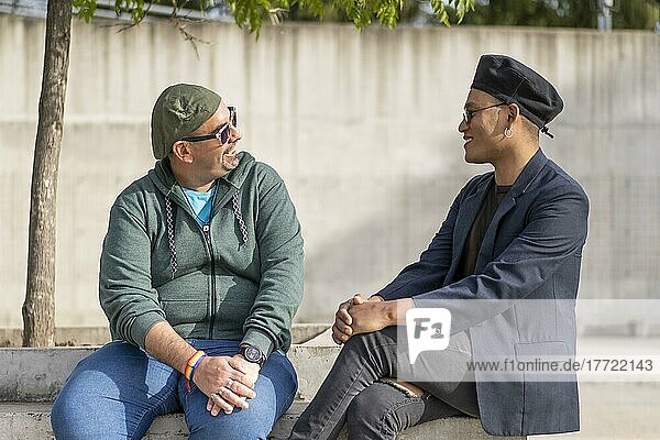 Gay Latino male couple sitting on a bench in a park  wearing fashionable hats and sunglasses  looking at each other smiling