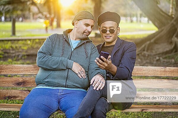 Gay Latino male couple sitting on a bench in a park at sunset  wearing fashionable hats  holding a cell phone  smiling